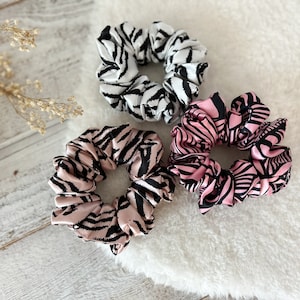 Puede incluir: Tres coleteros de satén con estampado de cebra. Uno es blanco y negro, otro rosa y negro, y el tercero rosa claro y negro. Estos accesorios para el cabello son una adición elegante a cualquier atuendo.