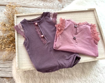 Op de afbeelding: Twee mouwloze baby rompertjes met kantversiering. Het rompertje links is donkerpaars met witte kant en roze knopen. Het rompertje rechts is lichtpaars met roze kant.