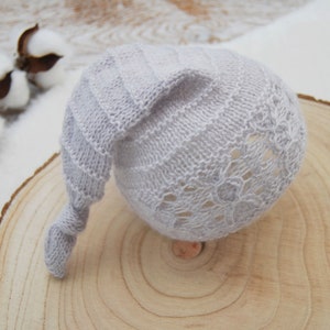 May include: A light gray knitted baby hat with a long, knotted tail. The hat has a delicate lace pattern and is perfect for newborn photography.