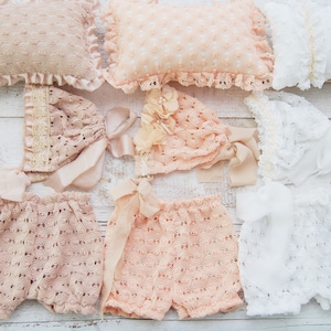 Könnte beinhalten: Ein Set gehäkelter Babykleidung in Weiß und Pfirsich, bestehend aus Mütze, Shorts und Oberteil. Die Kleidung liegt auf einer weißen Holzoberfläche.