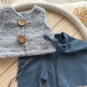 K&ouml;nnte beinhalten: Eine graue, flauschige Weste mit Holzkn&ouml;pfen und eine blaue, gemusterte Hose mit einem Krawattendetail.