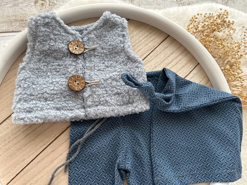 K&ouml;nnte beinhalten: Eine graue, flauschige Weste mit Holzkn&ouml;pfen und eine blaue, gemusterte Hose mit einem Krawattendetail.