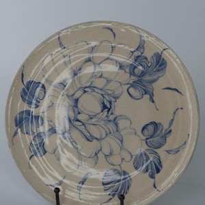 Puede incluir: Un plato redondo de cerámica con fondo beige y diseño floral azul. El plato presenta una flor grande en el centro con hojas circundantes. El plato se exhibe sobre un soporte de metal negro.