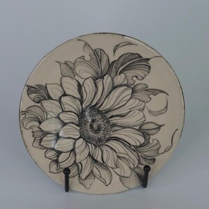 Puede incluir: Un plato decorativo redondo, de color crema, con un diseño floral detallado en blanco y negro. El plato está sostenido por un soporte de metal negro, que muestra la intrincada obra de arte de una gran flor con pétalos y hojas detallados.