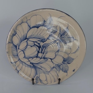 Puede incluir: Un plato redondo de cerámica con un diseño floral azul dibujado a mano. El plato tiene un fondo color crema y un borde azul oscuro. El diseño floral está centrado en el plato y presenta grandes pétalos estilizados.