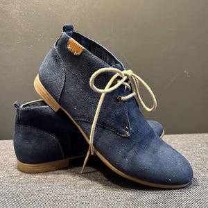 Op de afbeelding: Een paar marineblauwe suède enkellaarsjes met beige zolen en crèmekleurige veters. De laarzen hebben een klassiek chukka boot ontwerp met een klein merklabel. De laarzen staan op een grijze ondergrond.