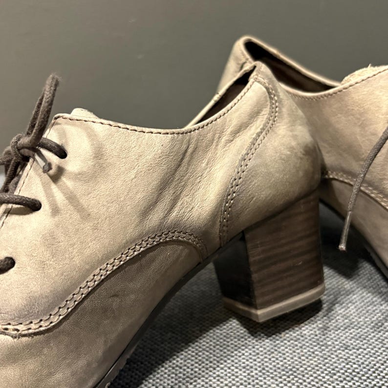 May include: Close-up of a pair of taupe-colored heeled oxford shoes. The shoes feature a lace-up design, a block heel, and visible stitching details. The leather appears worn, giving the shoes a vintage look.