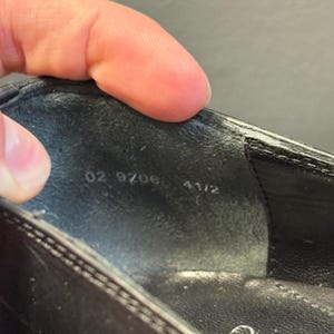 May include: Close-up of the inside of a black shoe. The shoe's interior is dark gray with the numbers "02 9706 4 1/2" printed in white. The shoe's exterior is a shiny black material.