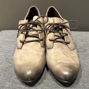May include: A pair of light gray leather oxford shoes with dark brown laces. The shoes have a low heel and a rounded toe. The leather has a distressed look, with darker shading on the toe and heel.