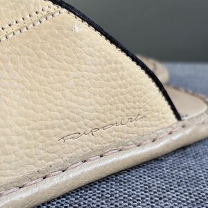 May include: A beige slide sandal featuring a textured, pebbled surface with visible stitching along the edge. The word Ripcurl is embossed in a cursive script on the side of the strap, which is accented by a thin black trim along the upper edge.