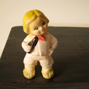 Può includere: Una piccola figurina di plastica di un bambino con i capelli biondi, che indossa una camicia bianca con una sciarpa rosso e bianca e tiene un oggetto marrone in mano.