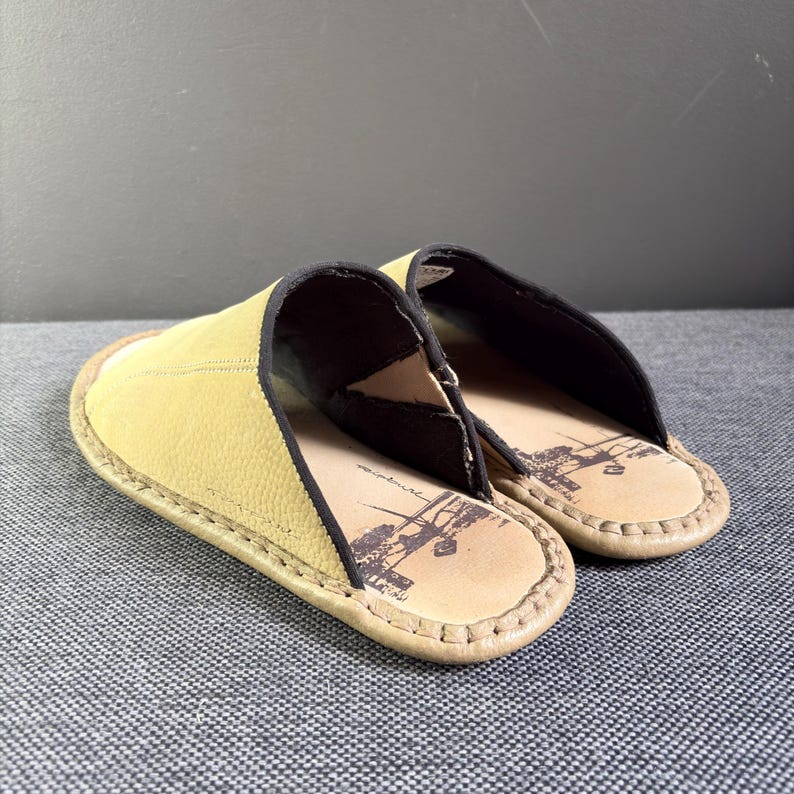 May include: A pair of slip-on slippers featuring a textured mustard yellow upper with black trim. The interior footbed is light tan and displays a dark brown graphic print of a vintage-style vehicle. The edges are finished with visible stitching.