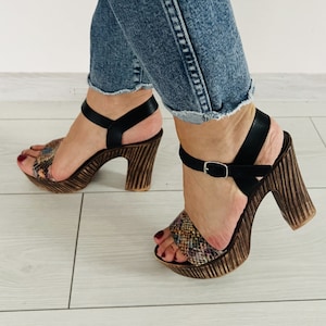 May include: A pair of brown wooden platform sandals with black straps and a snake print design.