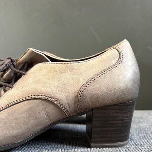May include: Close-up of a taupe-colored leather shoe with a block heel. The shoe features a lace-up design and visible stitching details. The heel is a dark brown color.