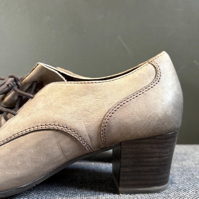 May include: Close-up of a taupe-colored leather shoe with a block heel. The shoe features a lace-up design and visible stitching details. The heel is a dark brown color.