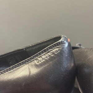 May include: Close-up of a pair of black leather shoes. The shoes have a stitched design along the top edge and the brand name "ANDYLL" is embossed on the side. A small red mark is visible on the heel.