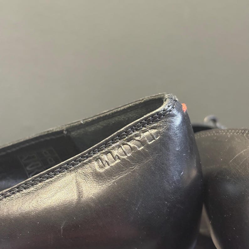 May include: Close-up of a pair of black leather shoes. The shoes have a stitched design along the top edge and the brand name "ANDYLL" is embossed on the side. A small red mark is visible on the heel.