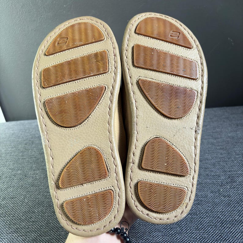 May include: A pair of tan shoes with light brown textured outsoles featuring a wavy pattern. The soles have a stitched border and a small logo embossed near the heel area. The shoes are positioned side by side, showing the bottom tread design.