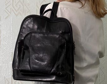 May include: A black leather backpack featuring a smooth, slightly glossy finish, a large main compartment, and a smaller front zippered pocket. The bag includes adjustable shoulder straps and a top handle, showcasing a structured design.