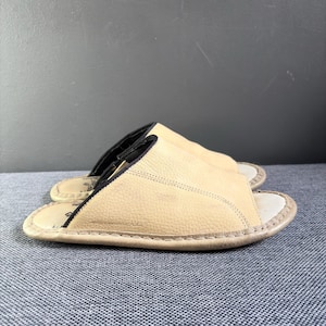 May include: A pair of beige leather slide sandals featuring a textured upper, an open-toe design, and a black interior lining. The footbed is light-colored with visible stitching along the perimeter and a small logo embossed on the side of the sole.