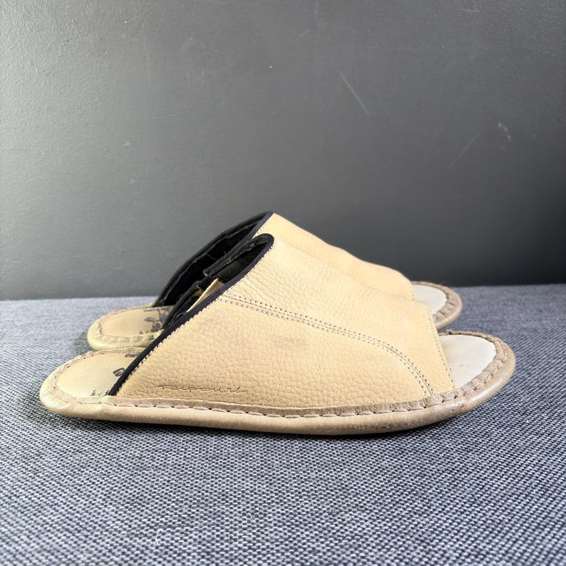 May include: A pair of beige leather slide sandals featuring a textured upper, an open-toe design, and a black interior lining. The footbed is light-colored with visible stitching along the perimeter and a small logo embossed on the side of the sole.