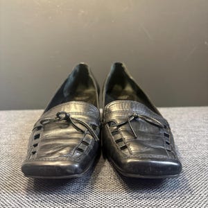 May include: A pair of black leather loafers with a square toe and decorative cutouts. Each shoe features a small bow detail on the vamp. The shoes are displayed on a gray surface.
