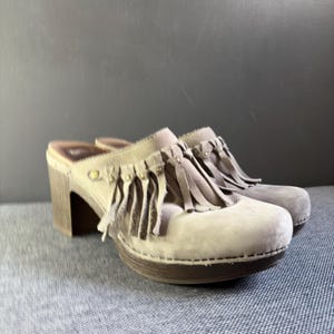 May include: A pair of beige suede clogs featuring a rounded toe, a block heel with a wood-grain texture, and a decorative fringe detail across the vamp secured with small metallic studs. The interior footbed is dark brown with visible branding.