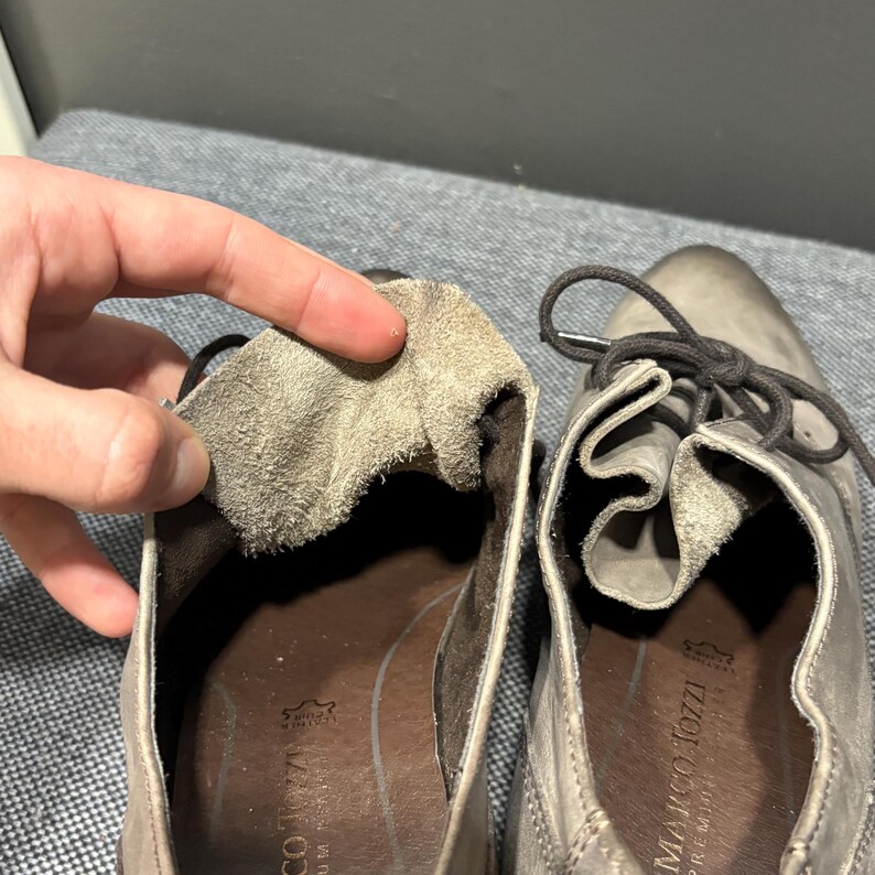 May include: Close-up of a pair of gray leather shoes with laces. The interior of one shoe is exposed, revealing a soft, textured lining. The brand name "MARCO TOZZI PREMIUM" is visible on the insole.