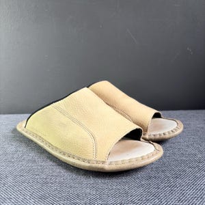 May include: A pair of beige open-toe slide sandals featuring a textured leather upper with visible stitching along the edges and a light-colored footbed. The design includes a black inner lining and a rounded sole, providing a simple slip-on style.