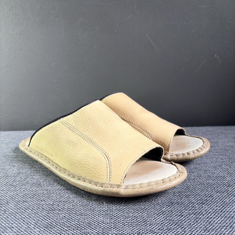 May include: A pair of beige open-toe slide sandals featuring a textured leather upper with visible stitching along the edges and a light-colored footbed. The design includes a black inner lining and a rounded sole, providing a simple slip-on style.