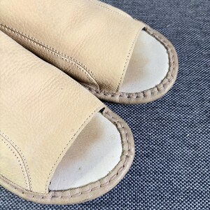 May include: A pair of open-toe slides featuring a textured beige leather upper and a smooth off-white footbed. The edges of the slides are finished with a thick, visible whipstitch detail in a matching beige tone, securing the upper to the sole.