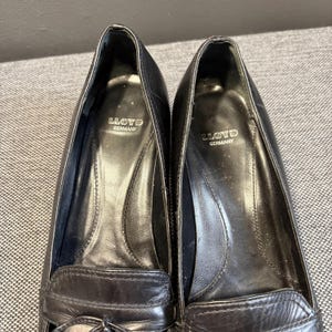May include: Close-up of two black leather loafers. The shoes have a classic design with a rounded toe and a decorative strap across the top. The brand name "LLOYD GERMANY" is visible inside each shoe.