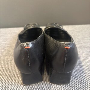 May include: A pair of black leather shoes with a low heel. The shoes have a rounded toe and a small bow detail on the top. The back of each shoe has a small orange stitch detail.