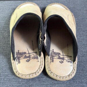 May include: A pair of beige slip-on sandals featuring a wide strap with black trim. The footbed is light beige and displays a dark brown graphic print of a surfer and the text Ripcurl. The edges of the sandals are finished with visible stitching.