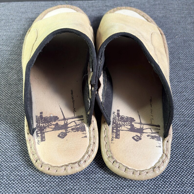 May include: A pair of beige slip-on sandals featuring a wide strap with black trim. The footbed is light beige and displays a dark brown graphic print of a surfer and the text Ripcurl. The edges of the sandals are finished with visible stitching.