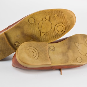 May include: A pair of brown leather shoes with a unique design on the soles. The soles feature a planet with rings and other circular designs.