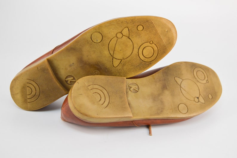 May include: A pair of brown leather shoes with a unique design on the soles. The soles feature a planet with rings and other circular designs.