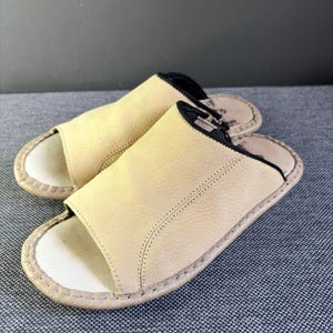 May include: A pair of beige open-toe slides featuring a textured leather upper with black trim along the edges. The footbed is light-colored with visible stitching around the perimeter of the sole, providing a simple and functional design for footwear.
