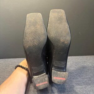 May include: A pair of black leather shoes with square toes. The soles are textured with a grid pattern. The heels have a red stripe. The shoes are held up by a person's hand.