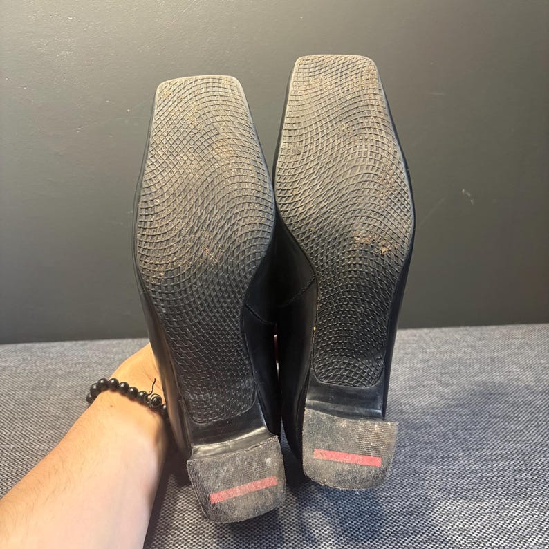 May include: A pair of black leather shoes with square toes. The soles are textured with a grid pattern. The heels have a red stripe. The shoes are held up by a person's hand.