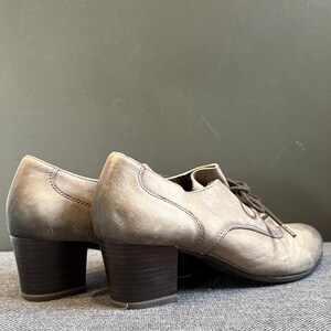 May include: A pair of gray leather heeled shoes with dark brown block heels and matching laces. The shoes have a classic oxford style with a rounded toe and visible stitching details.