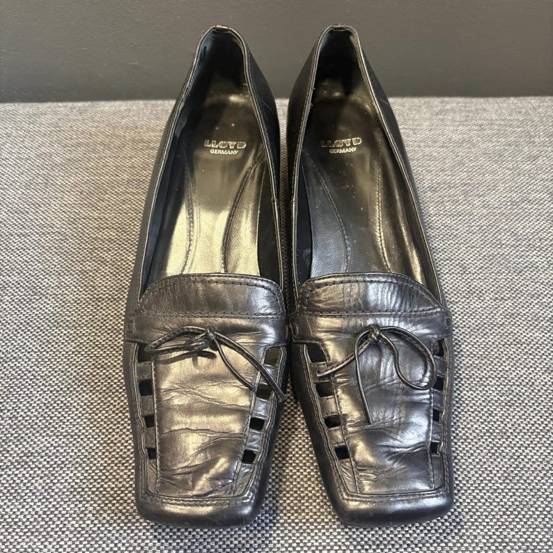 May include: A pair of black leather loafers with square toes. The shoes have a decorative cut-out design on the vamp and a small bow detail. The brand name "LLOYD GERMANY" is visible inside the shoe.