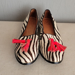 May include: A pair of black and white zebra print loafers with red tassels. The loafers have a pointed toe and a flat heel.