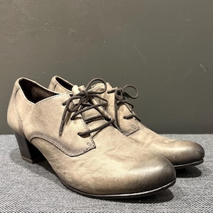 May include: A pair of gray leather oxford shoes with dark brown laces. The shoes have a rounded toe and a small, dark heel. The leather has a distressed appearance, giving them a vintage look. The shoes are on a gray surface.