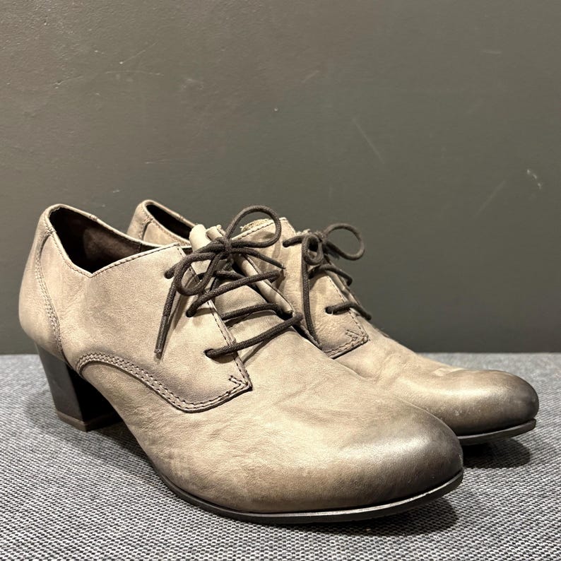 May include: A pair of gray leather oxford shoes with dark brown laces. The shoes have a rounded toe and a small, dark heel. The leather has a distressed appearance, giving them a vintage look. The shoes are on a gray surface.