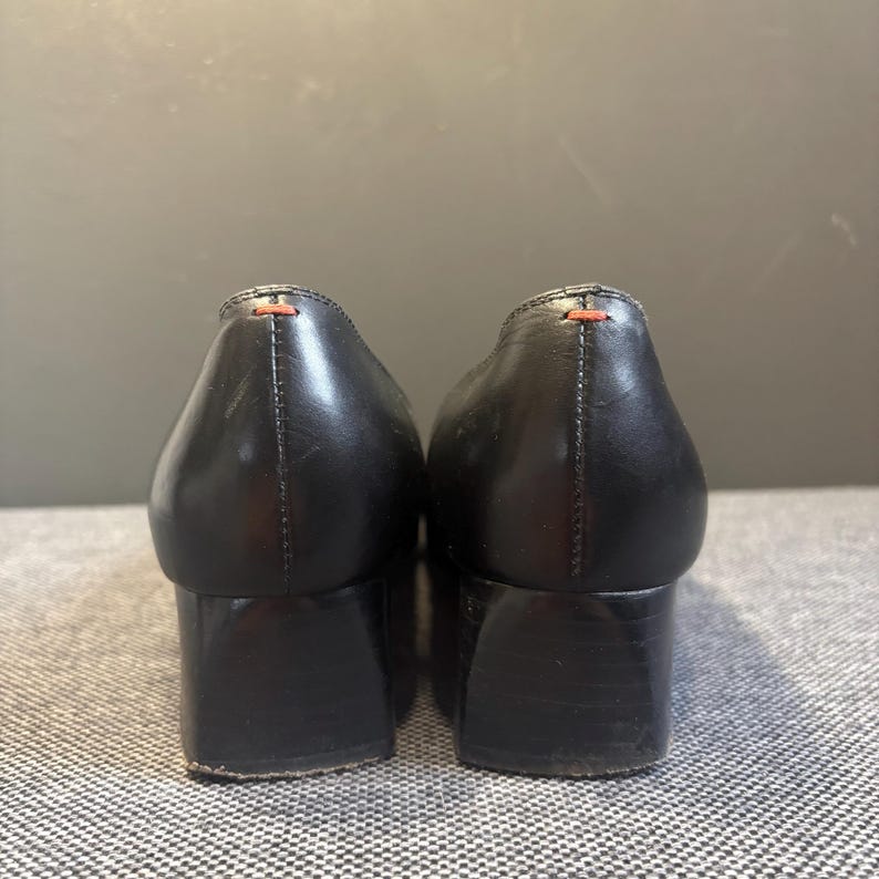 May include: A pair of black leather shoes with block heels. The shoes have a rounded toe and a small orange stitch detail at the heel. The shoes are set against a gray background.