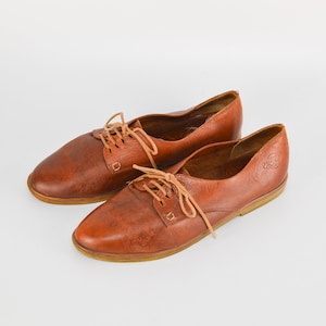 May include: A pair of brown leather lace-up shoes with a low heel. The shoes have a casual style and are made of a soft, supple leather.