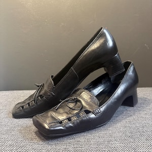 May include: A pair of black leather heeled loafers. The shoes have a square toe, cut-out details, and small bows. The heels are approximately 2 inches high.