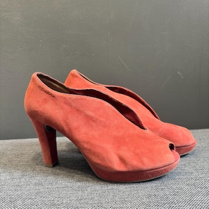 May include: A pair of coral-colored suede peep-toe pumps with a high heel and a platform sole. The shoes feature a deep V-shaped cutout at the vamp and the brand name Tamaris is embossed on the outer side of the heel area of the shoe.