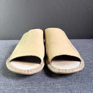 May include: A pair of open-toe slide sandals featuring a light tan, textured leather upper and a smooth, off-white footbed. The edges of the sandals are finished with visible stitching, and the interior lining appears to be a dark, contrasting color.